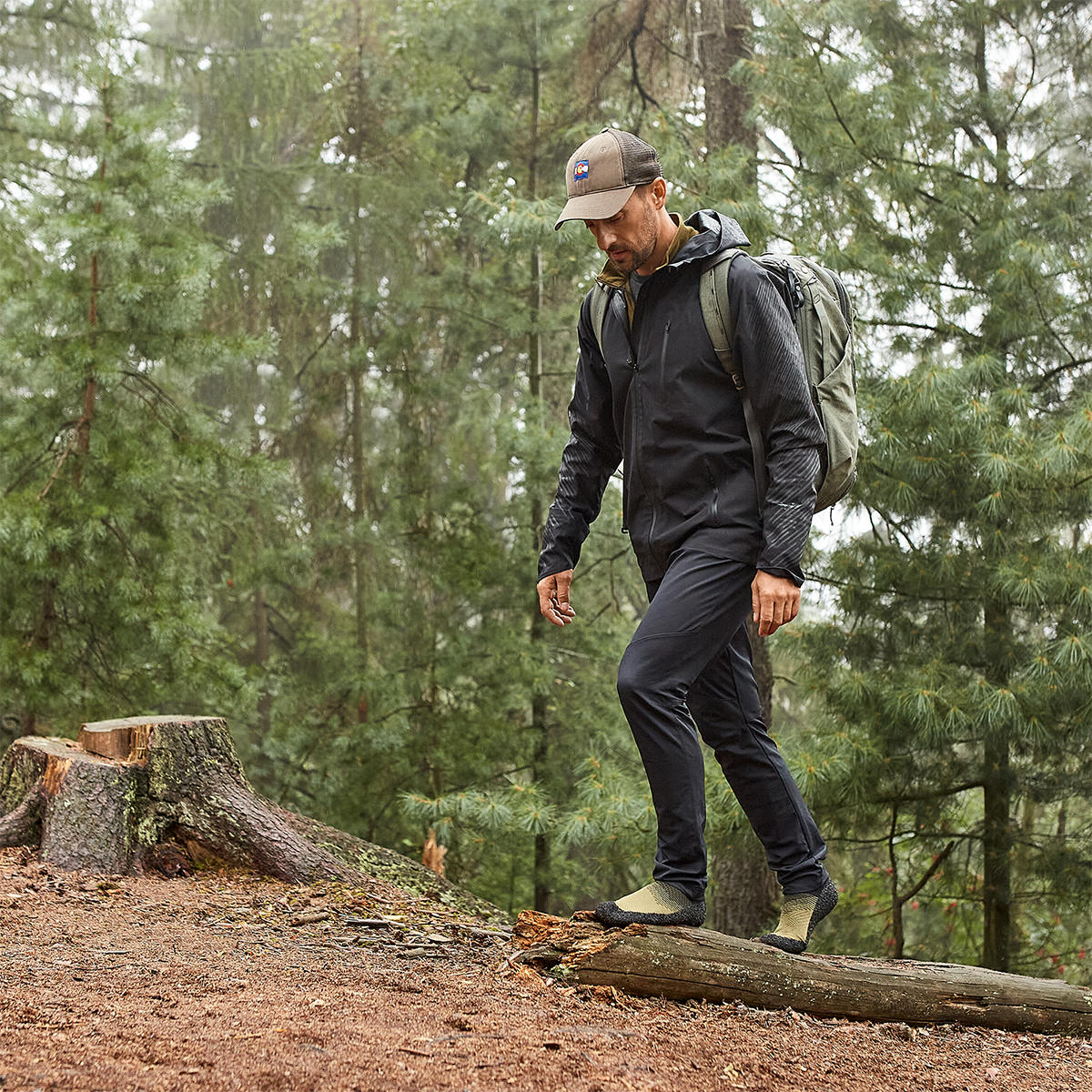 Person wearing Skinners sock shoes, walking across a wooden log in a forest.
