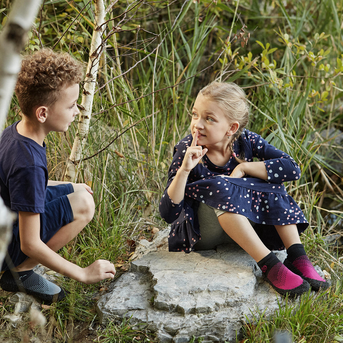 Two children sitting on a flat rock in a forested area wearing Skinners kids sock shoes